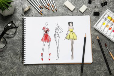 Gri desenli masadaki farklı kıyafetlerin çizimleri. Moda tasarımcısının masasında kırtasiye malzemesi var.
