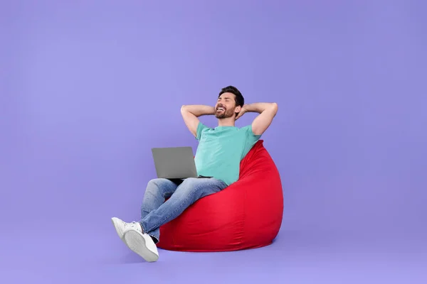 Homem Feliz Com Laptop Sentado Cadeira Beanbag Contra Fundo Roxo — Fotografia de Stock
