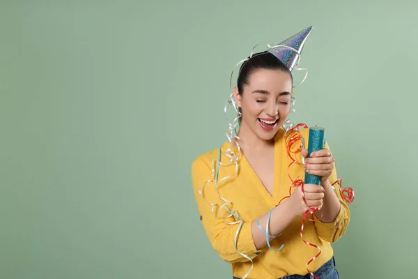 Young woman blowing up party popper on pale green background, space for text