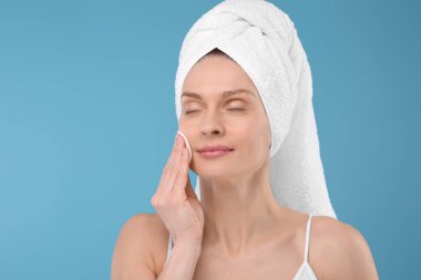 Terry havlusu içinde güzel bir kadın açık mavi arka planda pamuk yastıkla makyajı kaldırıyor.
