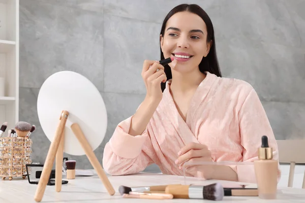 stock image Beautiful young woman applying lipstick at dressing table indoors