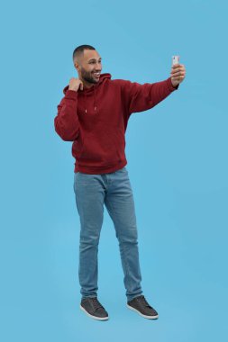 Gülümseyen genç adam açık mavi arka planda akıllı telefonuyla selfie çekiyor.