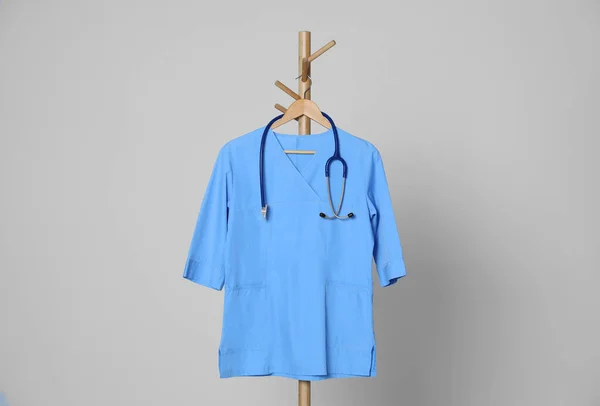 Medical uniform and stethoscope hanging on rack against light grey background