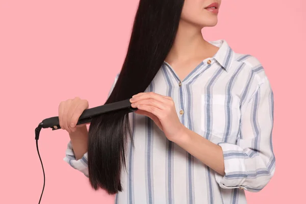 Woman using hair iron on pink background, closeup