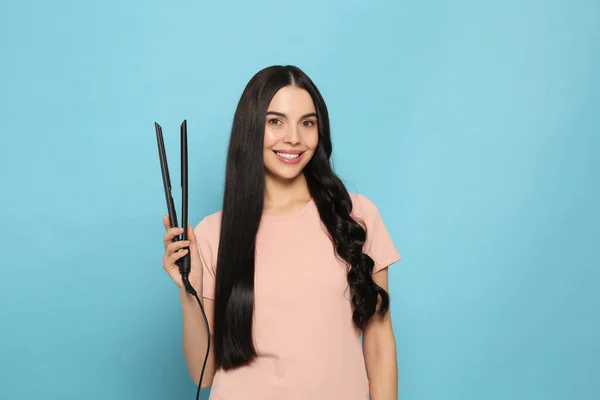 Beautiful happy woman with hair iron on light blue background
