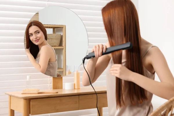 Beautiful woman using hair iron near mirror in room