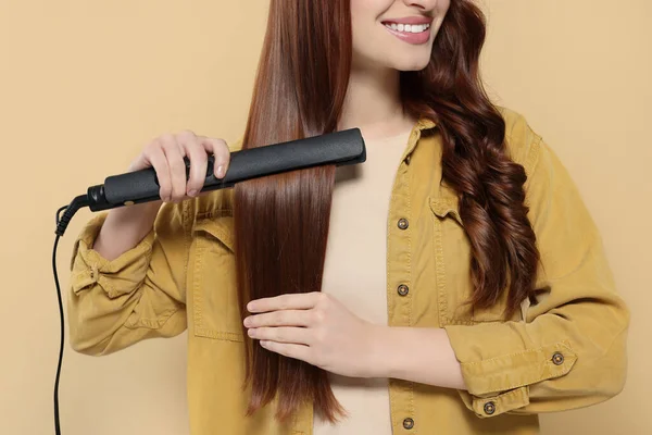 Young woman using hair iron on beige background, closeup