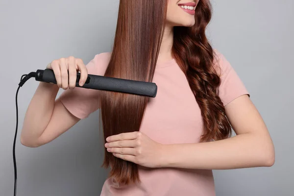 Young woman using hair iron on light gray background, closeup