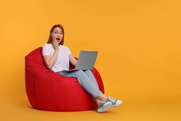 坐在黄色背景的豆袋椅上 坐在笔记本电脑上的令人惊讶的年轻女子 — 图库照片