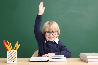 Küçük mutlu okul çocuğu masa başında karatahtanın yanında kitaplarla el kaldırıyor.