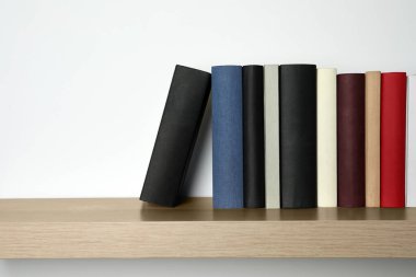 Beyaz duvarın yanındaki ahşap rafta bir sürü ciltli kitap var.