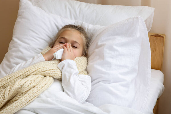Sick girl with scarf and tissue lying in bed while blowing nose indoors