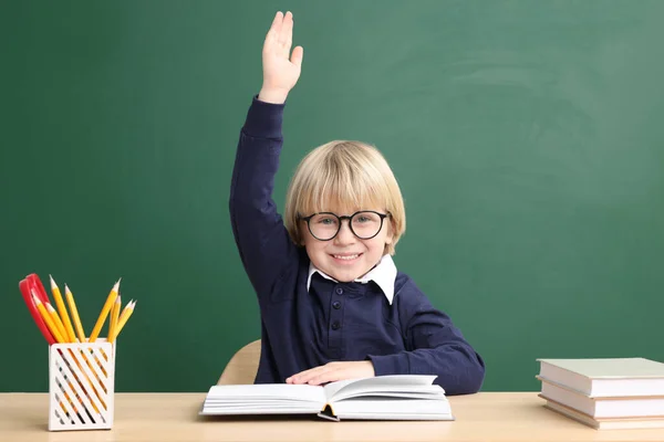 Счастливый Маленький Школьник Поднимает Руку Сидя Столом Книгами Возле Доски — стоковое фото