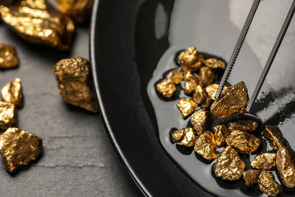 stock image Tweezers with gold nugget above pile on black table