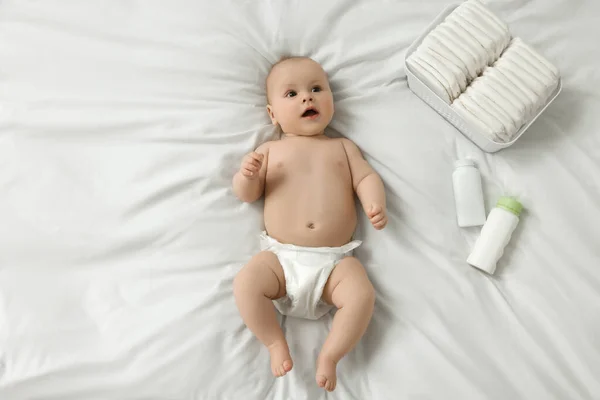 Cute baby, diapers and cosmetic products on white bed, top view