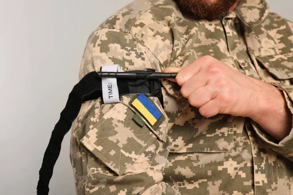 Ukrainian soldier in military uniform applying medical tourniquet on arm against light grey background, closeup