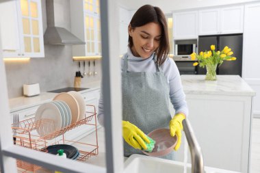Modern mutfakta lavabonun üstünde genç ve mutlu bir kadın bulaşık yıkıyor.