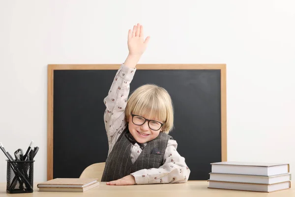 教室で黒板の近くの本と机に座っている間に手を上げる幸せな小さな学校の子供 — ストック写真