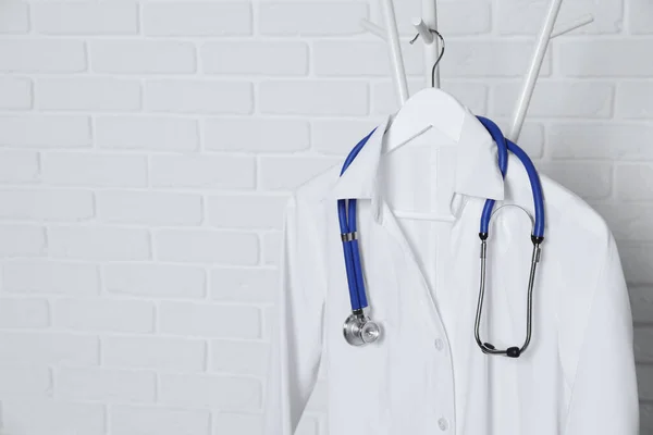 Medical uniform and stethoscope hanging on rack near white brick wall. Space for text