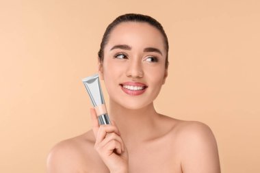 Bej arka planda fondöten tüpü tutan kadın.