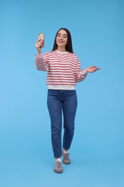 Gülümseyen genç bir kadın açık mavi arka planda akıllı telefonuyla selfie çekiyor.
