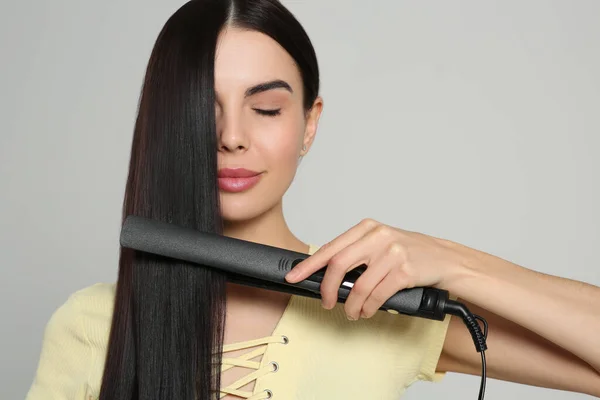 Beautiful woman using hair iron on light grey background