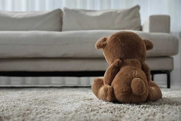 stock image Cute lonely teddy bear on floor near sofa in room, back view. Space for text