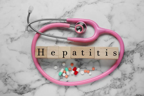 stock image Word Hepatitis made of wooden cubes, stethoscope and pills on white marble table, flat lay