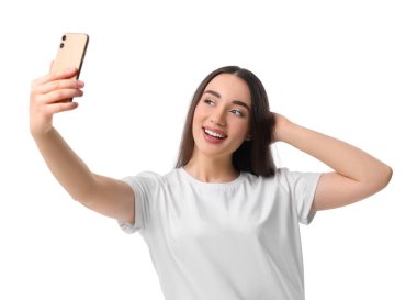 Gülümseyen genç bir kadın beyaz arka planda akıllı telefonuyla selfie çekiyor.