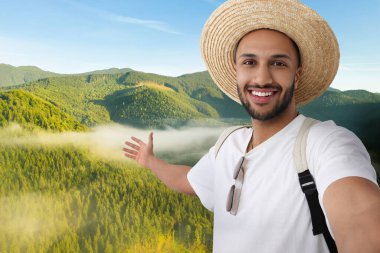 Hasır şapkalı gülümseyen genç adam dağlarda selfie çekiyor.