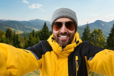 Şapkalı ve güneş gözlüklü gülen adam dağlarda selfie çekiyor.