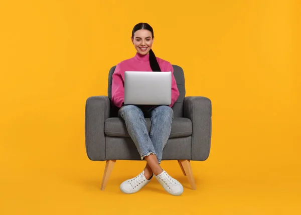 Happy woman with laptop sitting in armchair on orange background