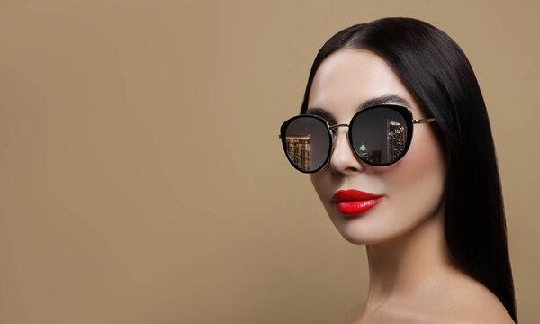Attractive woman in stylish sunglasses on dark beige background. Night cityscape reflecting in lenses. Space for text
