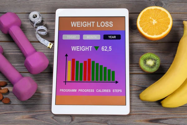 Tablet with weight loss calculator application, dumbbells and products on grey wooden table, above view