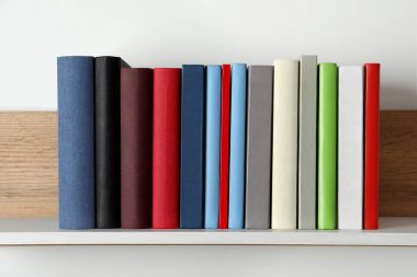 Beyaz duvarın yanındaki ahşap rafta bir sürü ciltli kitap var.
