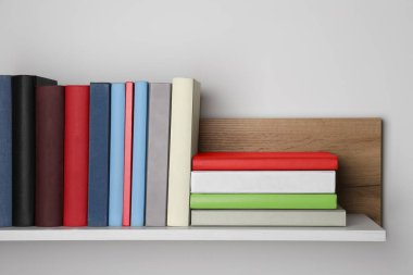 Beyaz duvarın yanındaki ahşap rafta bir sürü ciltli kitap var.