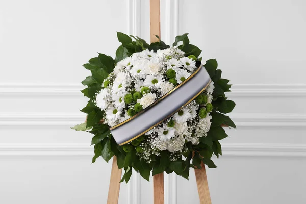 Funeral wreath of flowers with ribbon on wooden stand near white wall indoors