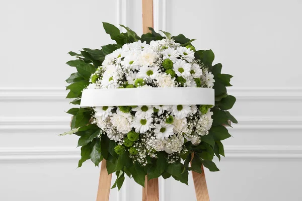 Funeral wreath of flowers with ribbon on wooden stand near white wall indoors