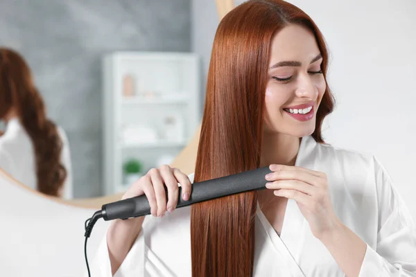 Beautiful woman using hair iron in room, space for text