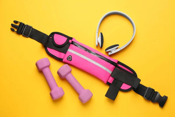 Stylish pink waist bag, dumbbells and headphones on yellow background, flat lay