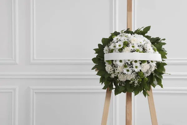 Funeral wreath of flowers with ribbon on wooden stand near white wall indoors, space for text