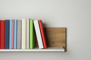 Beyaz duvarın yanındaki ahşap rafta bir sürü ciltli kitap var.