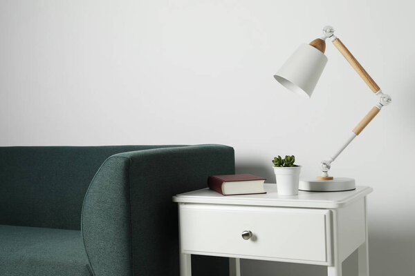 Stylish lamp, houseplant with book on side table and soft sofa near white wall. Interior design