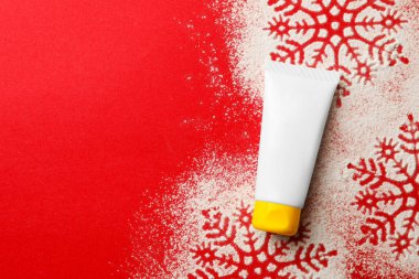 Winter skin care. Hand cream and snowflake silhouettes made with artificial snow on red background, top view. Space for text