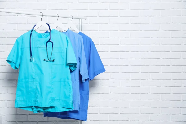 Medical uniforms and stethoscope hanging on rack near white brick wall. Space for text