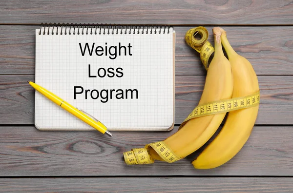 Notebook with phrase Weight Loss Program, measuring tape and bananas on grey wooden table, flat lay