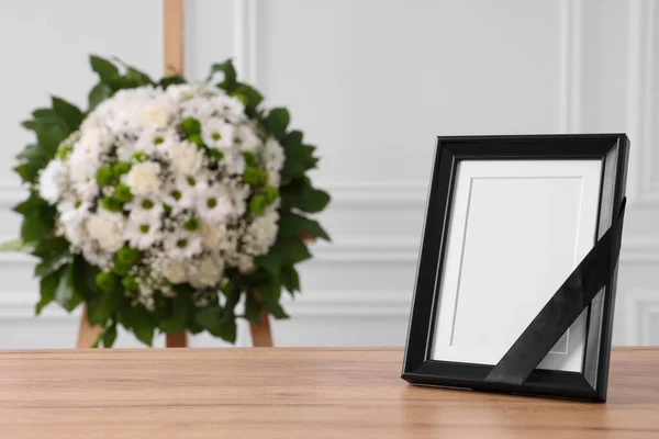 テーブルの上に黒いリボンと白い壁の近くの花の花輪を屋内で テキストのためのスペースを持つ写真フレーム 葬儀の属性 — ストック写真