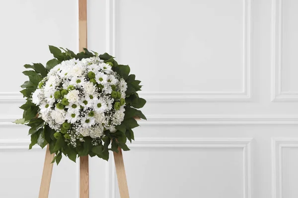 Funeral wreath of flowers on wooden stand near white wall indoors. Space for text