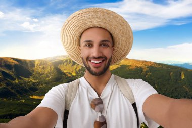 Hasır şapkalı gülümseyen genç adam dağlarda selfie çekiyor.