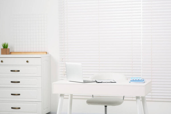Beautiful office with laptop on white table, chair and chest of drawers. Doctor's workplace